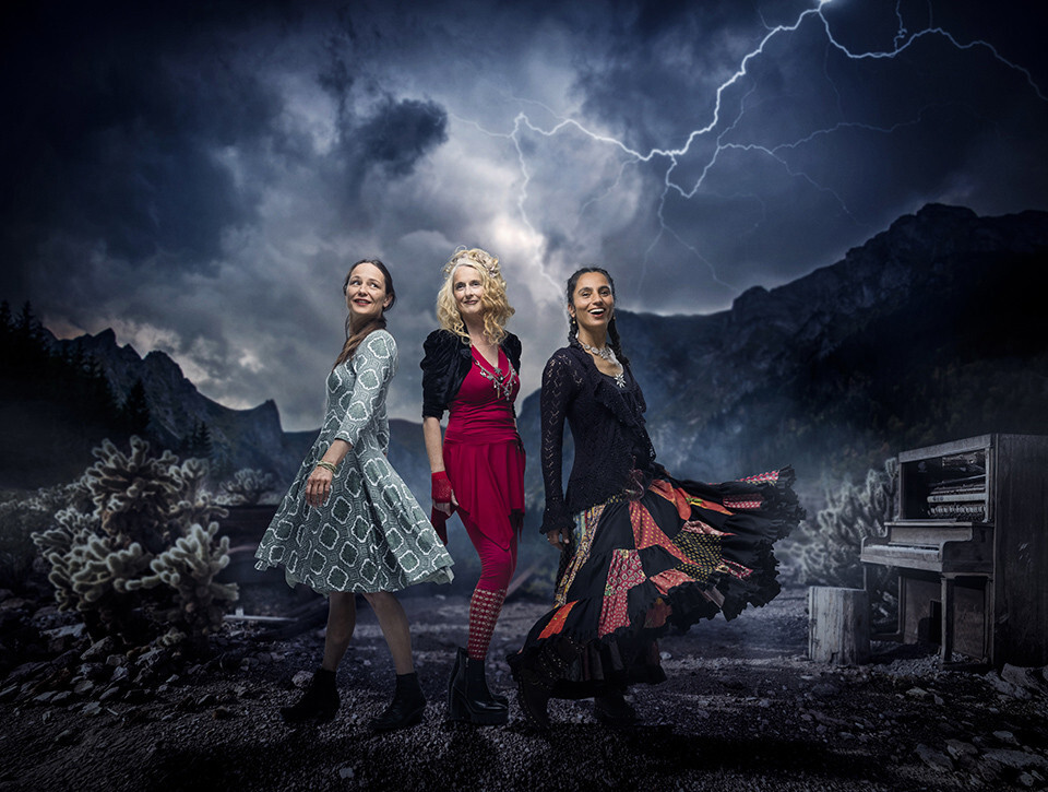 Die Rote Zora mit Barbara Berger, Christine Lauterburg und Natalie Huber in farbenfrohen Kleidern vor einer dramatischen Berglandschaft mit Gewitterhimmel, zum Auftritt im Schlosskeller Interlaken.