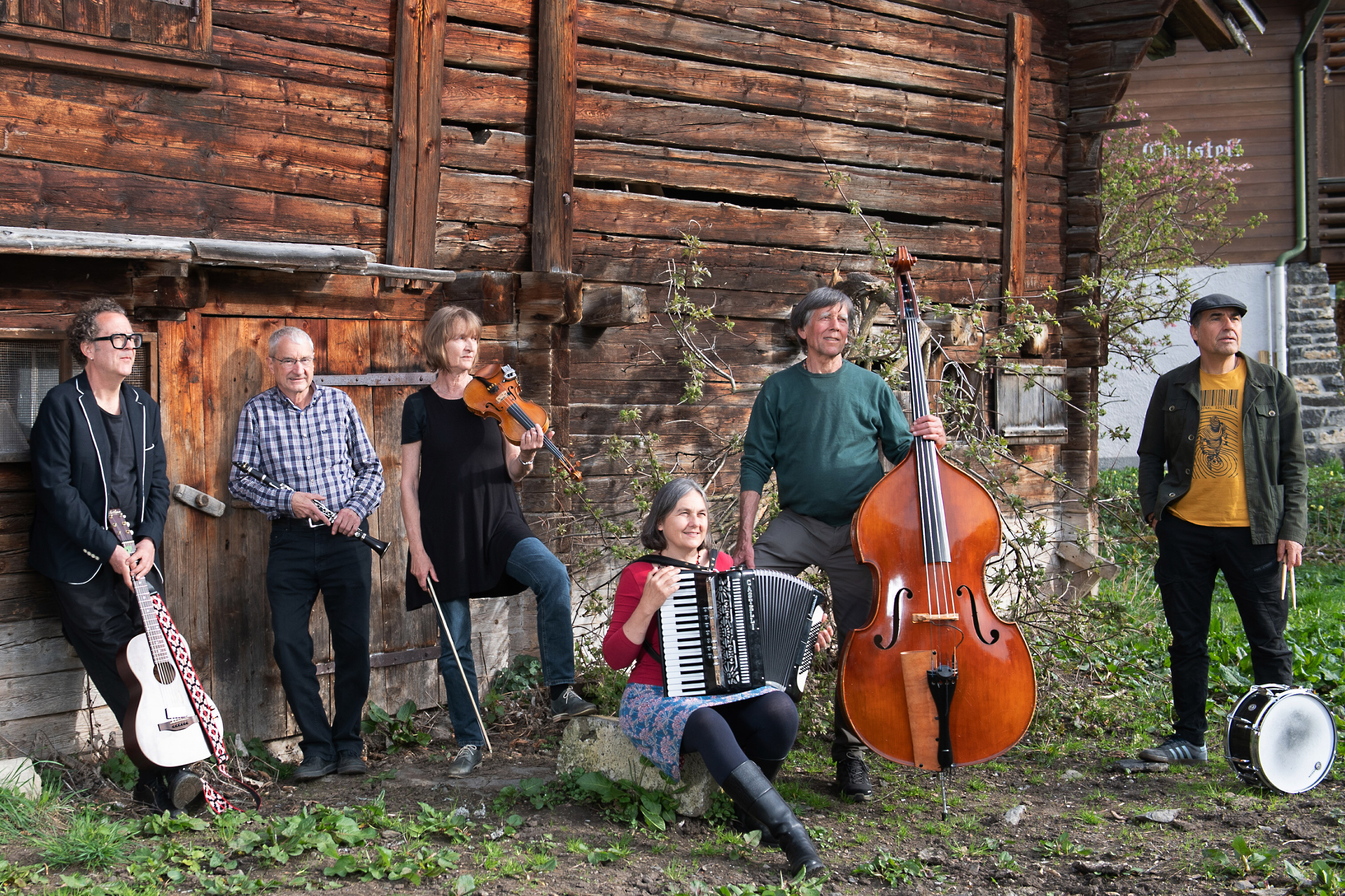 Permis-B, Klänge, Melodien und Lieder aus fremden Kulturen werden heimisch, die Musiker in einer Reihe vor einem Holzhaus, zum Auftritt im Schlosskeller Interlaken.